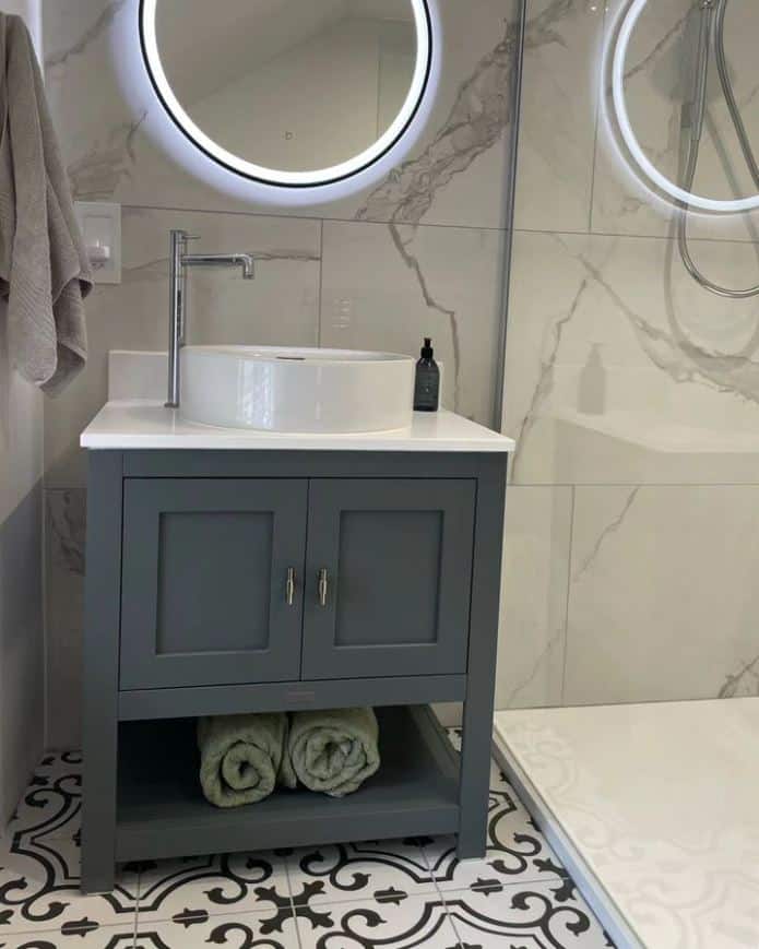 A painted vanity unit in dark grey with two doors, a round sink sit on top of a white countertop with chrome tap and chrome handles, towels sit under the unit on a shelf